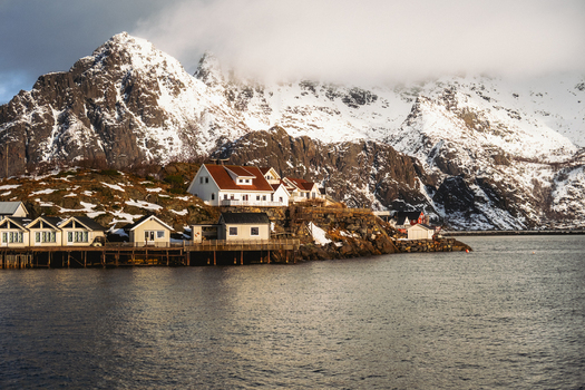 View of white houses with red roofs nestled along the coast contrasting with the snow capped mountain backdrop and calm waters Henningsv&aelig;r Nordland Norway.