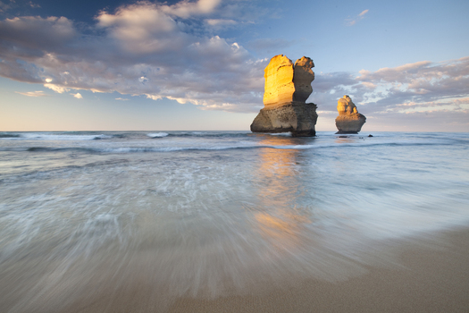 View of the limestone stacks of the Twelve Apostles Marine National Park with waves washing onto the sandy beach under a cloudy sky at sunset Australia. View of the limestone stacks of the Twelve Apostles Marine National Park with waves washing onto the sandy beach under a cloudy sky at sunset Australia.
