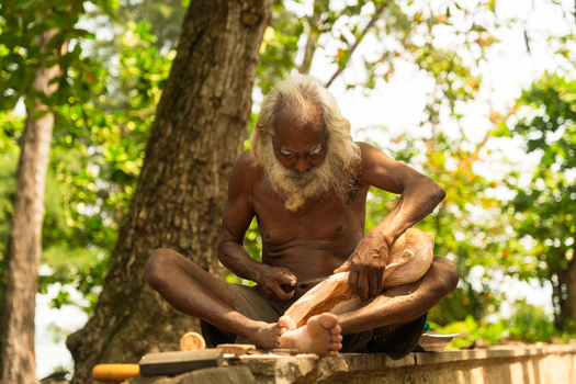 Sri Lanka 12 February 2026 View of an elderly man with a long white beard carving a wooden sculpture while sitting outdoors under lush green trees.