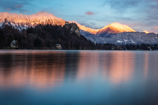 View of Lake Bled and Bled Castle with snow capped mountains illuminated by golden sunset light and calm water reflections Bled Radovljica Slovenia. View of Lake Bled and Bled Castle with snow capped mountains illuminated by golden sunset light and calm water reflections Bled Radovljica Slovenia.