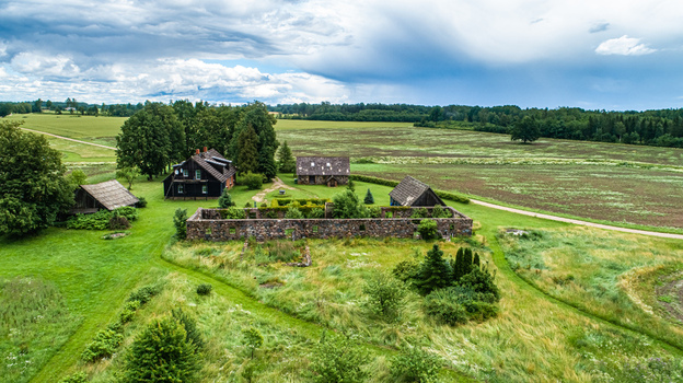 Aerial view of a historic stone ruin and a traditional wooden house surrounded by lush green fields and trees in the rural countryside of Latvia. Aerial view of a historic stone ruin and a traditional wooden house surrounded by lush green fields and trees in the rural countryside of Latvia.