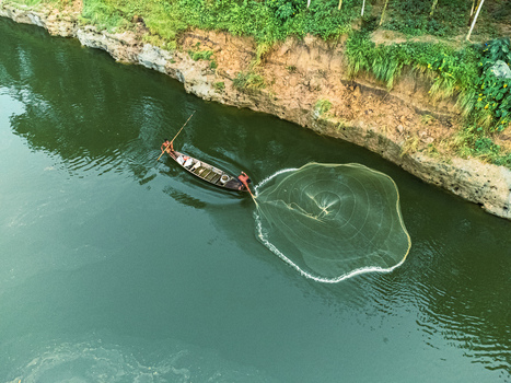 Khansama Upazila Bangladesh 12 September 2023 Aerial view of fishermen in a small wooden boat casting a circular net into the green river water near a lush riverbank.