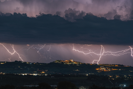 View of the hilltop town of Montecosaro illuminated at night under a dramatic thunderstorm with multiple lightning strikes across the dark sky in Montecosaro Marche Italy. View of the hilltop town of Montecosaro illuminated at night under a dramatic thunderstorm with multiple lightning strikes across the dark sky in Montecosaro Marche Italy.