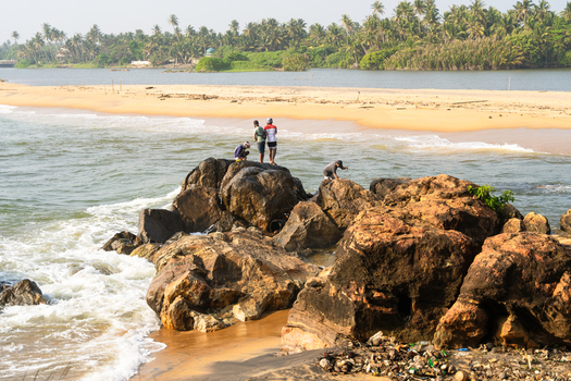 Sri Lanka 21 January 2026 View of people standing on large coastal rocks as waves crash against the shore with a sandy beach and palm trees in the background.