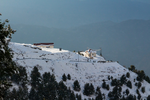 Aerial view of snow dusted buildings and pathways atop a hill contrasting sharply with the muted greens of the distant mountains Pir Chanasi Azad Kashmir Pakistan.