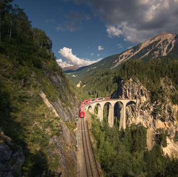 Aerial view of the Landwasser Viaduct with a red train crossing the stone arches in a steep mountain valley surrounded by pine trees in Bergun Filisur Graubunden Switzerland.