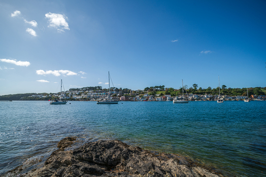 View of the Fowey River with sailboats anchored near the historic town of Fowey featuring colorful buildings along the waterfront and a rocky foreground in Fowey Cornwall United Kingdom. View of the Fowey River with sailboats anchored near the historic town of Fowey featuring colorful buildings along the waterfront and a rocky foreground in Fowey Cornwall United Kingdom.