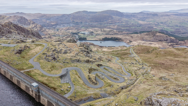 Aerial view of the winding road snaking through the rugged terrain near the dam and lake framed by distant mountains Blaenau Ffestiniog Wales United Kingdom. Aerial view of the winding road snaking through the rugged terrain near the dam and lake framed by distant mountains Blaenau Ffestiniog Wales United Kingdom.