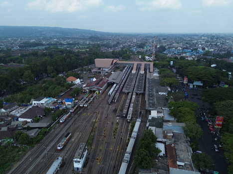Aerial view of Stasiun Malang Kota Baru with trains parked at the platforms surrounded by urban buildings and lush trees in Kecamatan Klojen Jawa Timur Indonesia.