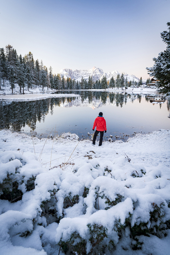 View of a solitary figure in a vibrant red jacket standing at the snow covered edge of a tranquil lake reflecting the sky and distant mountains Misurina Veneto Italy. View of a solitary figure in a vibrant red jacket standing at the snow covered edge of a tranquil lake reflecting the sky and distant mountains Misurina Veneto Italy.