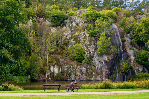 Ronneby Blekinge County Sweden 11 September 2025 View of Brunssparken featuring a waterfall on a rocky cliff with two people on a bench by the pond in the historic park. Ronneby Blekinge County Sweden 11 September 2025 View of Brunssparken featuring a waterfall on a rocky cliff with two people on a bench by the pond in the historic park.
