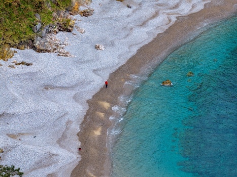Aerial view of Silence Beach Viewpoint with a person in a red jacket walking on the pebble shore by turquoise waters and green cliffs Cudillero Asturias Spain.