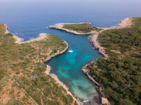Aerial view of Cala Sa Nau bay with turquoise water a white sailboat and rocky cliffs covered in green vegetation in Felanitx Balearic Islands Spain. Aerial view of Cala Sa Nau bay with turquoise water a white sailboat and rocky cliffs covered in green vegetation in Felanitx Balearic Islands Spain.