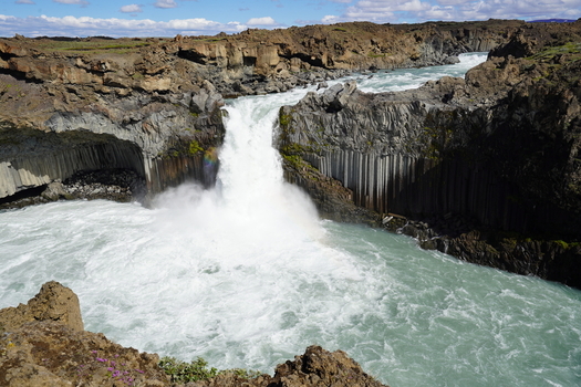 View of Aldeyjarfoss waterfall cascading over basalt columns into a turquoise pool surrounded by rugged volcanic rock formations under a bright blue sky in Iceland. View of Aldeyjarfoss waterfall cascading over basalt columns into a turquoise pool surrounded by rugged volcanic rock formations under a bright blue sky in Iceland.