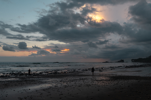 View of people walking along a rocky beach at sunset with dramatic storm clouds and golden light over the ocean in Costa Rica.