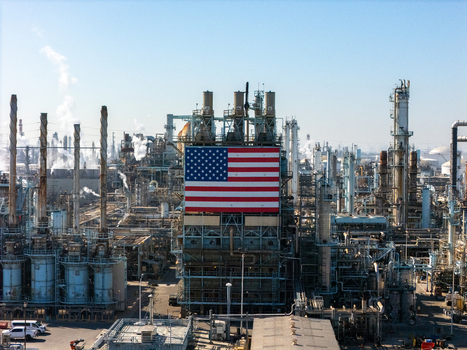 Aerial view of an oil refinery with a large American flag on a central structure surrounded by industrial piping storage tanks and smokestacks. Carson California United States.