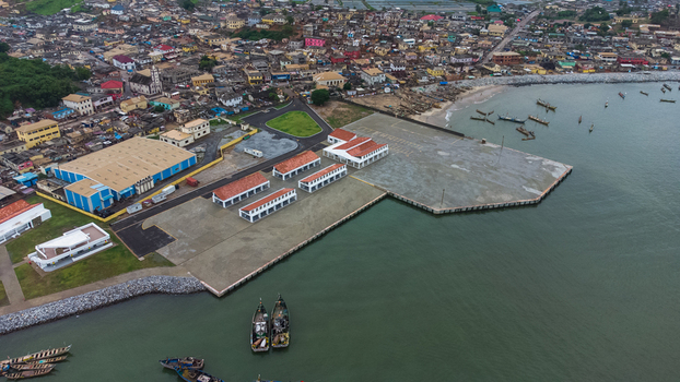 Aerial view of the modern harbor facilities and fishing port in Elmina with traditional boats moored in the water and the historic coastal town in Elmina Central Region Ghana. Aerial view of the modern harbor facilities and fishing port in Elmina with traditional boats moored in the water and the historic coastal town in Elmina Central Region Ghana.