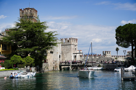 View of the Scaligero Castle with its medieval towers and stone walls overlooking the canal with boats and tourists in Sirmione Lombardy Italy.
