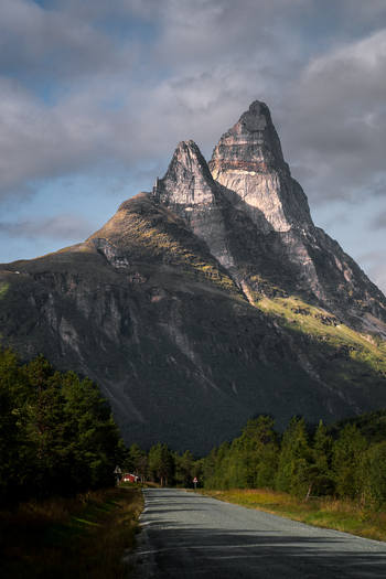 View of Otertinden mountain rising above a quiet road lined with pine trees under a dramatic cloudy sky in Storfjord Troms Norway. View of Otertinden mountain rising above a quiet road lined with pine trees under a dramatic cloudy sky in Storfjord Troms Norway.