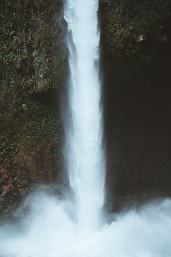 View of a powerful waterfall cascading down a mossy dark rock cliff surrounded by lush tropical vegetation in Costa Rica.