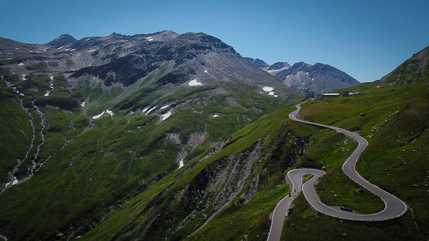 Aerial view of the winding hairpin turns of the Furka Pass mountain road surrounded by green alpine slopes and rocky peaks in Obergoms Valais Switzerland. Aerial view of the winding hairpin turns of the Furka Pass mountain road surrounded by green alpine slopes and rocky peaks in Obergoms Valais Switzerland.