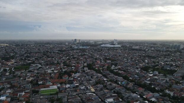 Aerial view of the dense urban landscape featuring residential rooftops and a prominent stadium in the distance under a cloudy sky in Bekasi Barat West Java Indonesia. Aerial view of the dense urban landscape featuring residential rooftops and a prominent stadium in the distance under a cloudy sky in Bekasi Barat West Java Indonesia.