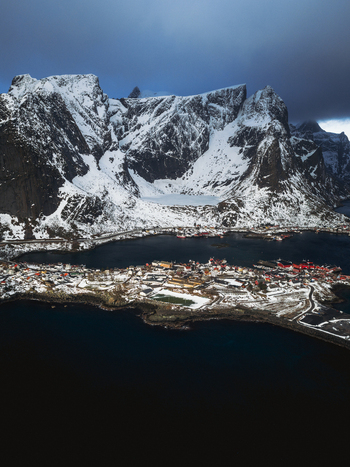 Aerial view of jagged snow dusted peaks meet the tranquil dark waters surrounding a quaint village dotted with red roofed houses Reinebringen Nordland Norway.