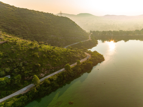 Aerial view of a winding road along the edge of a lake surrounded by lush green hills under the warm glow of the setting sun in Udaipur Rajasthan India. Aerial view of a winding road along the edge of a lake surrounded by lush green hills under the warm glow of the setting sun in Udaipur Rajasthan India.