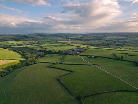 Aerial view of the rolling green agricultural landscape featuring patchwork fields traditional hedgerows farm tracks and a distant wind turbine in Looe Cornwall United Kingdom. Aerial view of the rolling green agricultural landscape featuring patchwork fields traditional hedgerows farm tracks and a distant wind turbine in Looe Cornwall United Kingdom.