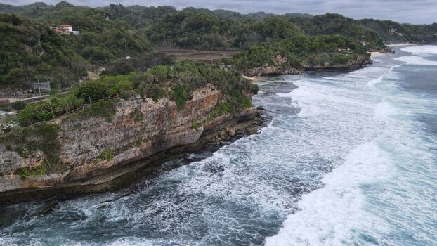 Aerial view of Indrayanti Beach with rugged limestone cliffs crashing waves and lush tropical vegetation along the coastline in Kecamatan Tepus Daerah Istimewa Yogya Indonesia. Aerial view of Indrayanti Beach with rugged limestone cliffs crashing waves and lush tropical vegetation along the coastline in Kecamatan Tepus Daerah Istimewa Yogya Indonesia.
