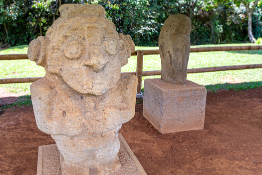 View of San Agustin Archaeological Park featuring ancient pre Columbian stone statues standing on pedestals in a lush outdoor setting with a bamboo fence in San Agustin Huila Colombia.
