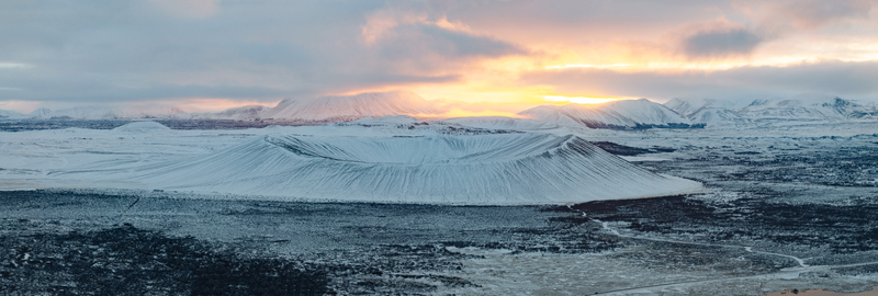 Aerial view of Hverfjall crater covered in snow under a golden sunset sky with distant mountains in Thingeyjarsveit Iceland. Aerial view of Hverfjall crater covered in snow under a golden sunset sky with distant mountains in Thingeyjarsveit Iceland.