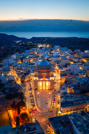 Aerial view of the Parish Church of the Assumption of the Blessed Virgin Mary into Heaven illuminated at dusk surrounded by the town of Mgarr Regjun Tramuntana Malta.