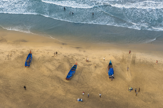 Aerial view of traditional wooden fishing boats resting on the sandy beach with people walking along the shore and swimmers in the ocean waves in Kovalam Keralam India. Aerial view of traditional wooden fishing boats resting on the sandy beach with people walking along the shore and swimmers in the ocean waves in Kovalam Keralam India.