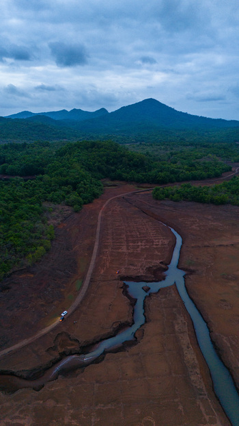 Aerial view of the exposed red earth landscape and winding stream of the submerged village of Kurdi surrounded by lush green hills under a cloudy sky in Kurdi Goa India.
