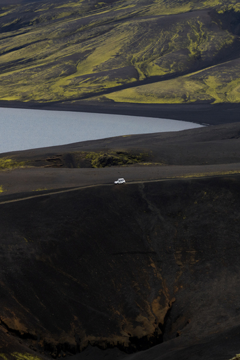 View of a lone white vehicle traverses a stark black volcanic landscape contrasted by a serene lake and green hills Skaftárhreppur Iceland. View of a lone white vehicle traverses a stark black volcanic landscape contrasted by a serene lake and green hills Skaftárhreppur Iceland.