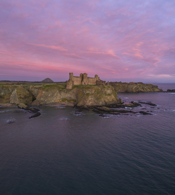Aerial view of the ruined Tantallon Castle perched on high cliffs overlooking the dark sea under a vibrant pink sunset sky in North Berwick Scotland United Kingdom.