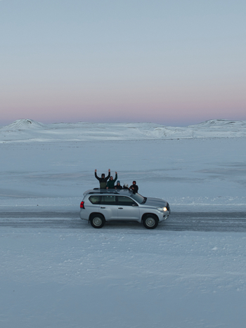 Aerial view of a silver SUV driving through a vast snow covered landscape with people standing on the roof under a soft twilight sky in Kirkjubaejarklaustur Skaftarhreppur Iceland. Aerial view of a silver SUV driving through a vast snow covered landscape with people standing on the roof under a soft twilight sky in Kirkjubaejarklaustur Skaftarhreppur Iceland.