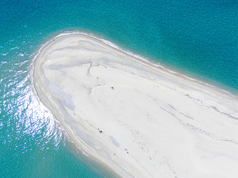 Aerial view of Possidi Beach sand spit surrounded by crystal clear turquoise waters and deep blue sea with people on the white sand in Greece.