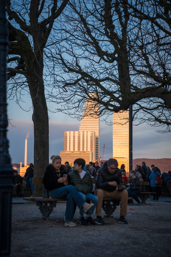 Basel Switzerland 10 March 2025 View of people enjoying the evening light on a bench with the stark geometric architecture of the BIZ tower rising in the background. Basel Switzerland 10 March 2025 View of people enjoying the evening light on a bench with the stark geometric architecture of the BIZ tower rising in the background.