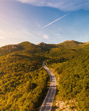 Aerial view of a winding road through lush green forested hills under a clear blue sky with a vapor trail during golden hour Rijeka Primorje Gorski Kotar Croatia. Aerial view of a winding road through lush green forested hills under a clear blue sky with a vapor trail during golden hour Rijeka Primorje Gorski Kotar Croatia.