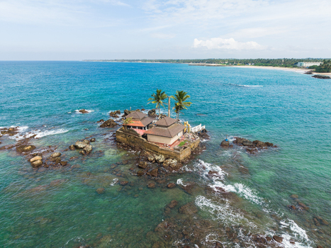 Aerial view of Seenigama Muhudu Dewalaya temple on a rocky outcrop surrounded by turquoise ocean waves and palm trees Peraliya Sri Lanka. Aerial view of Seenigama Muhudu Dewalaya temple on a rocky outcrop surrounded by turquoise ocean waves and palm trees Peraliya Sri Lanka.