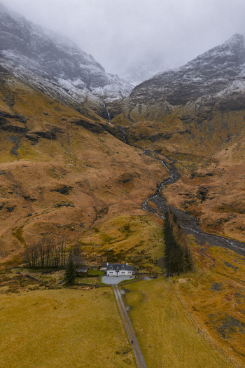 Aerial view of the iconic white cottage nestled at the base of snow dusted mountains with a winding stream and rugged terrain Glencoe Scotland United Kingdom. Aerial view of the iconic white cottage nestled at the base of snow dusted mountains with a winding stream and rugged terrain Glencoe Scotland United Kingdom.