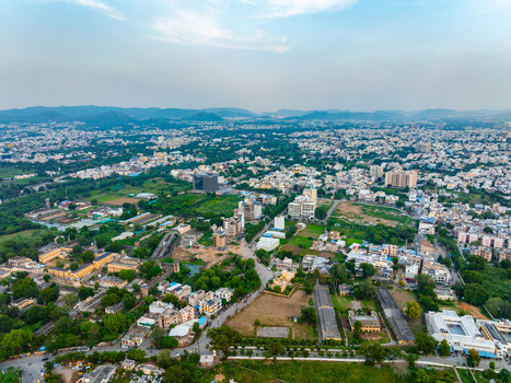 Aerial view of the sprawling urban landscape featuring residential buildings lush green patches and distant hills under a clear sky in Udaipur Rajasthan India. Aerial view of the sprawling urban landscape featuring residential buildings lush green patches and distant hills under a clear sky in Udaipur Rajasthan India.
