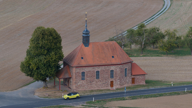 Aerial view of Wendelinuskapelle surrounded by agricultural fields and trees with a yellow car parked on the road in Monchberg Bayern Germany. Aerial view of Wendelinuskapelle surrounded by agricultural fields and trees with a yellow car parked on the road in Monchberg Bayern Germany.