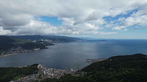 Aerial view of the coastal city of Atami nestled between lush green mountains and the deep blue waters of Sagami Bay under a cloudy sky in Atami Shizuoka Japan.