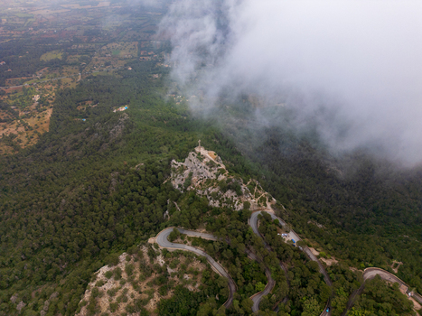 Aerial view of the Sanctuary of Sant Salvador cross on a rocky peak above winding mountain roads and dense pine forests under a misty sky Felanitx Illes Balears Spain. Aerial view of the Sanctuary of Sant Salvador cross on a rocky peak above winding mountain roads and dense pine forests under a misty sky Felanitx Illes Balears Spain.