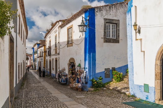 Obidos Portugal 13 May 2026 View of historic streets in Obidos with traditional white washed buildings featuring blue trim cobblestone pavement and a shop display under a clear sky. Obidos Portugal 13 May 2026 View of historic streets in Obidos with traditional white washed buildings featuring blue trim cobblestone pavement and a shop display under a clear sky.