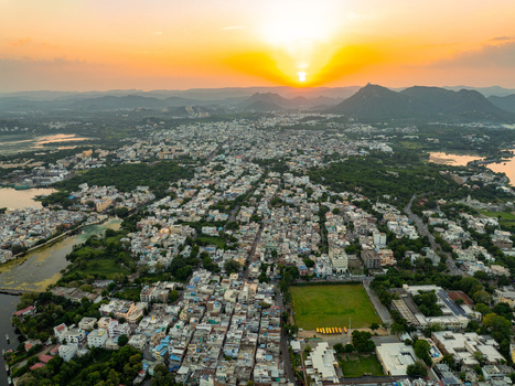 Aerial view of the city of Udaipur at sunset featuring dense urban architecture surrounding lakes and distant hills under a golden sky in Udaipur Rajasthan India.