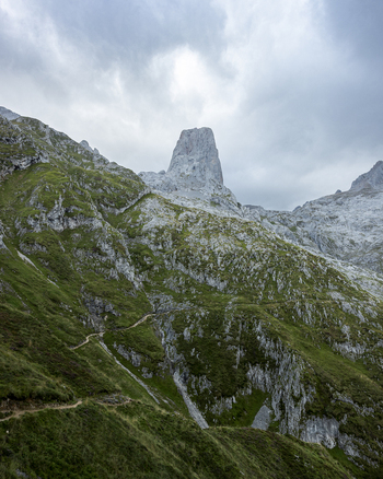 View of a jagged mountain peak piercing the sky amidst green slopes and winding trails under a cloudy sky Picos de Europa Asturias Spain. View of a jagged mountain peak piercing the sky amidst green slopes and winding trails under a cloudy sky Picos de Europa Asturias Spain.